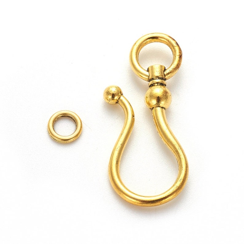 May include: Gold-toned metal watch chain hook with a round ring at the top and a curved hook at the bottom. The hook has a small round bead in the middle.