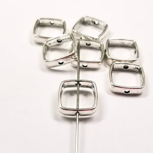 Puede incluir: Cuentas cuadradas plateadas con esquinas redondeadas. Cada cuenta tiene un pequeño agujero en lados opuestos y un acabado ligeramente envejecido. Las cuentas se muestran en una varilla de metal sobre un fondo blanco, adecuadas para la fabricación de joyas o manualidades.