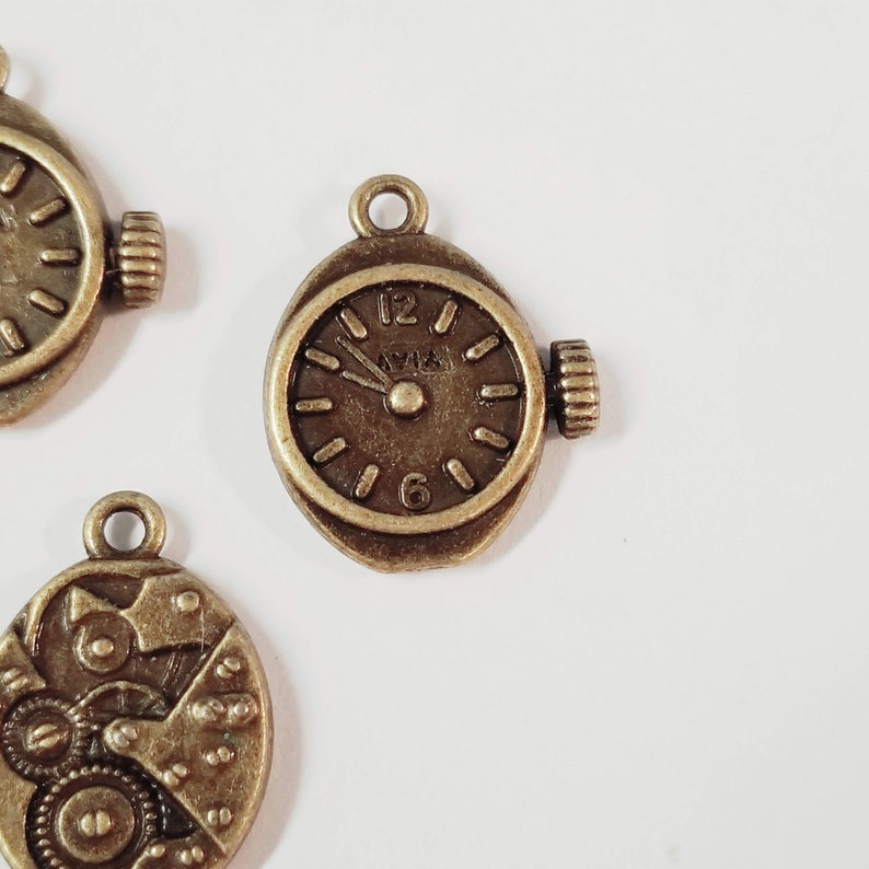 May include: A bronze-colored metal charm in the shape of a pocket watch with a clock face and Roman numeral markings. The charm has a loop at the top for attaching to a necklace or bracelet.