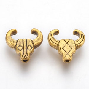 May include: Two gold-toned metal bull head charms with a geometric design. The charms have a hole at the bottom for attaching to jewelry or other crafts.