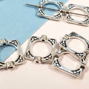 May include: Silver square-shaped metal beads with intricate designs. The beads have a hole in the center and are designed to be strung on a cord or chain.