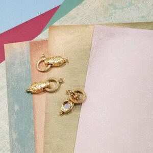 May include: Three gold-colored magnetic clasps for jewelry making. The clasps have an ornate design and a circular ring for attachment. The clasps are shown on a background of various colored papers.