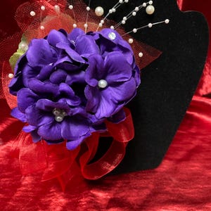 May include: A purple flower corsage with white pearl accents and a red ribbon. The corsage is displayed on a black velvet stand.