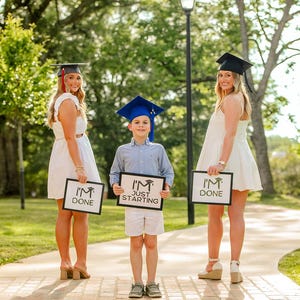 Może przedstawiać: Trzy osoby w czapkach absolwentów i białych sukienkach stoją na ceglanej ścieżce. Jedna osoba trzyma znak z napisem "I'M JUST STARTING", a pozostałe dwie znaki z napisem "I'M DONE". Scena rozgrywa się na zewnątrz z drzewami w tle.