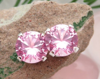 May include: A pair of pink round stud earrings with silver settings. The earrings are on a rock with a green moss-like growth.