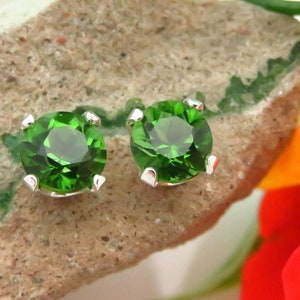 May include: A pair of silver stud earrings with round, green gemstones. The earrings are set against a background of a rock and a flower.