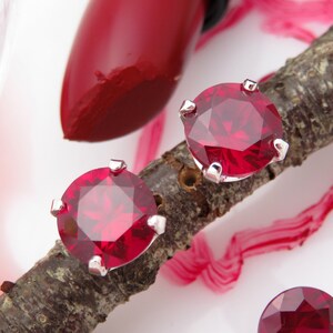 May include: A pair of silver stud earrings with red gemstone accents. The earrings are displayed on a wooden branch with a red lipstick tube and a red gemstone in the background.