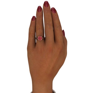 Puede incluir: Un anillo de oro rosa con una piedra preciosa rosa rodeada de diamantes m&aacute;s peque&ntilde;os. El anillo est&aacute; en una mano con esmalte de u&ntilde;as rojo.