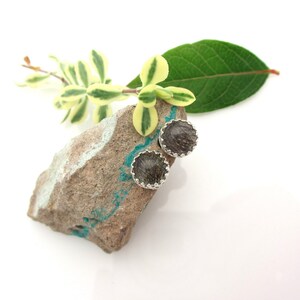 May include: A pair of silver stud earrings with a round, dark grey stone set in a silver bezel. The stones have a sparkly, iridescent quality.