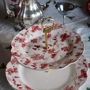 May include: Two-tiered serving tray with a gold handle. The white plates feature a red toile pattern and scalloped edges. The tray is designed for serving desserts or appetizers.