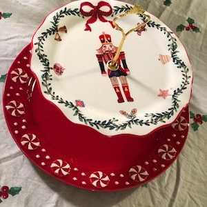 May include: A two-tiered Christmas serving platter. The bottom tier is red with white candy cane accents. The top tier is white with a red bow and a nutcracker soldier design. A gold handle connects the two tiers. Festive holiday decor.