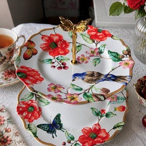 May include: A two-tier porcelain serving stand featuring white plates with scalloped gold edges. The design includes vibrant red flowers, green leaves, butterflies, and a blue bird. A gold-colored metal handle with a butterfly topper connects the tiers.
