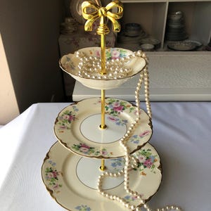 May include: A three-tiered serving stand with floral patterned plates and a gold-colored bow-shaped handle. A pearl necklace drapes over the plates. The plates have a cream-colored background with pink, blue, and green floral designs.