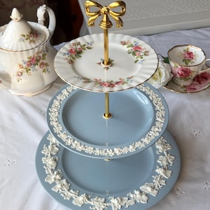 May include: A three-tier serving stand featuring a top white porcelain plate with floral patterns and two larger light blue plates with white embossed grape vine relief designs. The stand has a gold-colored metal central rod topped with a bow.
