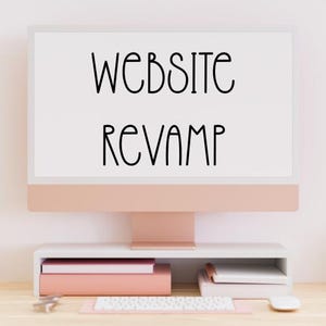 May include: A rose gold desktop computer monitor displaying the black text WEBSITE REVAMP on a white screen. The monitor sits on a white shelf above a white keyboard and mouse, with stacked pink and white books placed underneath the monitor screen.