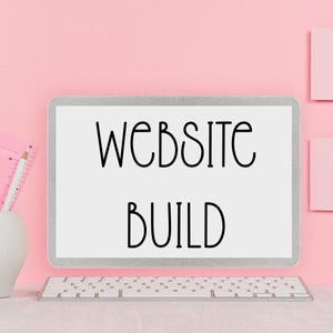 May include: A computer monitor displays the words "WEBSITE BUILD" in black against a white background. A white keyboard sits below the monitor. A white vase with pencils and a pink notepad are on the left, all against a pink backdrop.