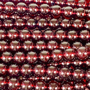 May include: Close-up of strands of polished, round, dark red garnet beads. The beads are strung together, creating a textured surface. The beads have a high-gloss finish, reflecting light. The overall effect is a rich, luxurious appearance.