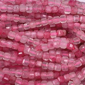 May include: Close-up of a collection of pink gemstone beads. The beads are square-shaped with faceted edges and a translucent appearance, strung together on a pink thread. The color varies from light to deep pink.