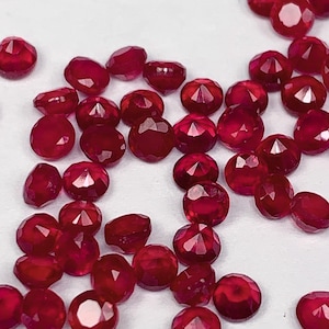 Puede incluir: Una colección de piedras preciosas de color rojo rubí, facetadas y pulidas, esparcidas sobre una superficie blanca. Las piedras preciosas son de varios tamaños y formas, con un color rojo intenso.