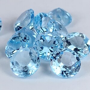 Pode incluir: Um conjunto de pedras preciosas azuis claras facetadas. As gemas, de forma redonda e oval, apresentam múltiplas facetas que capturam a luz. As pedras estão espalhadas sobre uma superfície branca, criando um efeito brilhante.