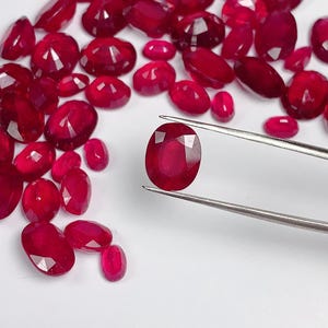 May include: A collection of loose, oval-cut red rubies with faceted surfaces. One single ruby is held by a pair of silver-toned metal tweezers, showcasing its deep red color and reflective cut against a plain white background.