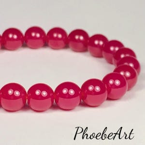 May include: A close-up of a bracelet featuring a strand of glossy, round, deep pink beads. The beads are evenly spaced and reflect light, creating small white highlights. The bracelet is set against a soft, white background. The text "PhoebeArt" is in the bottom right corner.