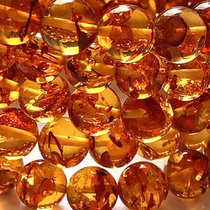 May include: Close-up of a collection of amber-colored, spherical beads. The beads have a glossy, reflective surface, with internal inclusions visible. The arrangement creates a textured, layered effect, with light reflecting off the surfaces.
