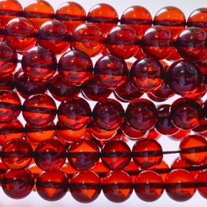 May include: A collection of polished, spherical beads in a deep, translucent reddish-orange hue. The beads are arranged in multiple rows, showcasing a smooth, glossy surface that reflects light, creating bright highlights across each individual sphere.