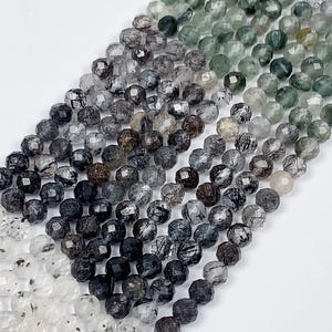 May include: A strand of faceted black tourmaline beads with inclusions. The beads are arranged in a gradient from light to dark, with the lightest beads at the bottom and the darkest beads at the top.