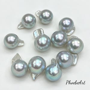 May include: A collection of ten loose, round, light gray pearls with iridescent surfaces and small, flat, irregular protrusions at the top of each piece. The text PhoebeArt is printed in black in the bottom right corner of the image.