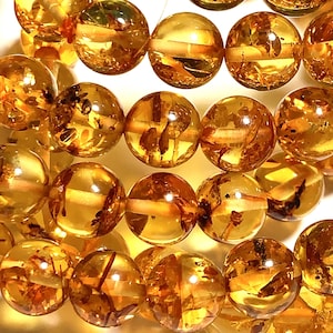 May include: Close-up of a collection of polished amber beads. The beads are round and translucent, with a warm, golden-yellow hue. Some beads have visible inclusions, adding to their unique appearance. The beads are strung together, suggesting they are for jewelry making.