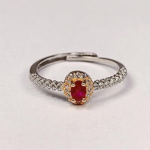 May include: A silver ring with a red ruby gemstone surrounded by smaller white gemstones. The ring has a textured band.