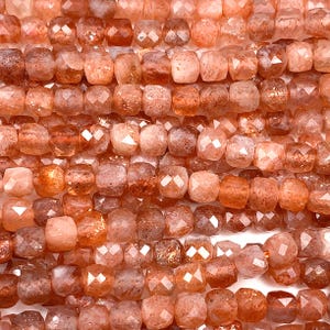 May include: A strand of faceted, orange sunstone beads. The beads are square shaped and have a shimmery, metallic finish.