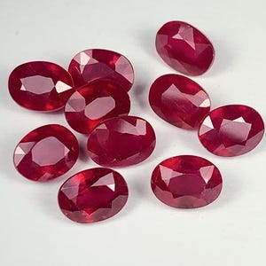 May include: A collection of oval-cut, deep red gemstones, likely rubies, are scattered on a white surface. The gems exhibit a high degree of clarity and a polished, reflective surface, showcasing their rich color and internal structure. The arrangement suggests a display of loose stones.