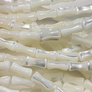 May include: Close-up of strands of ivory-colored, bone-shaped beads. The beads have a pearlescent sheen, reflecting light with a subtle iridescence. The beads are strung together, creating a textured and layered effect, suitable for jewelry making.