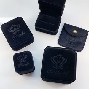 Puede incluir: Cuatro cajas de joyer&iacute;a de terciopelo negro con el logotipo "Phoebe" o "Phoebe Art" en relieve en la parte superior. Las cajas son de diferentes tama&ntilde;os y formas.