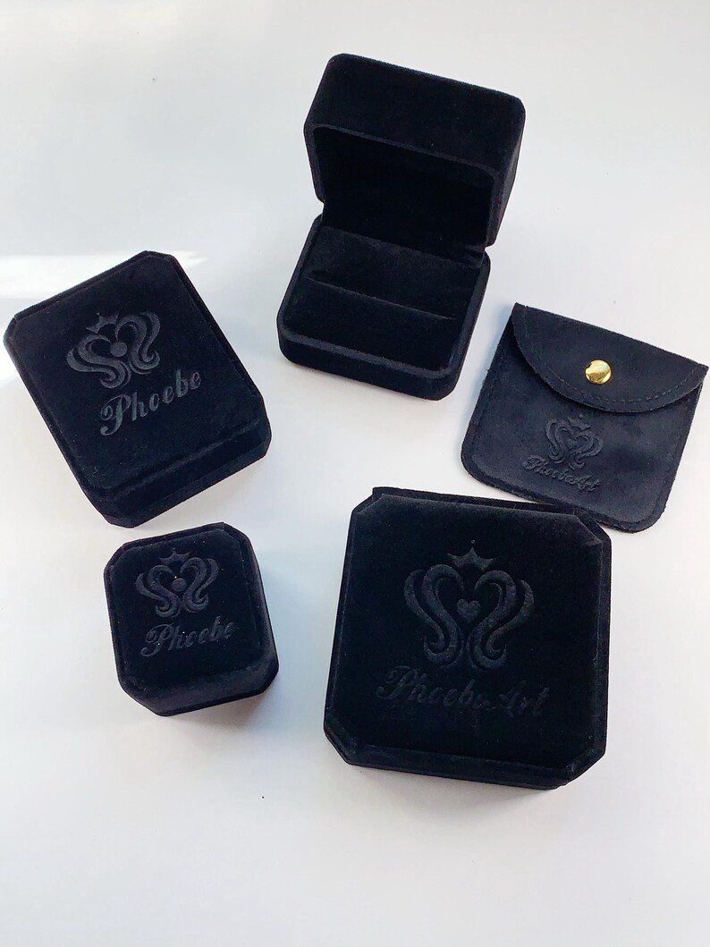 Puede incluir: Cuatro cajas de joyer&iacute;a de terciopelo negro con el logotipo "Phoebe" o "Phoebe Art" en relieve en la parte superior. Las cajas son de diferentes tama&ntilde;os y formas.