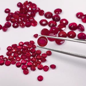 May include: A close-up of a pile of loose red gemstones, with one gemstone held by tweezers. The gemstones are round and have a faceted cut.