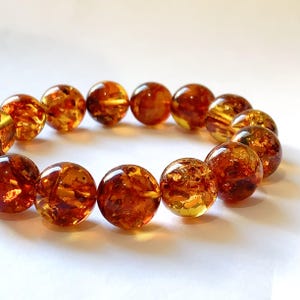May include: A bracelet composed of round, polished amber beads. Each bead displays a translucent, deep orange and golden-brown hue with visible internal inclusions and natural variations, arranged in a circular formation on a plain surface.