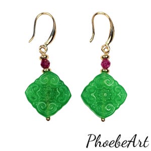 May include: A pair of gold-colored dangle earrings. Each earring features a bright green, carved jade pendant with a decorative design. A small, round, red bead and a gold bead are between the jade and the gold hook. The earrings are on a white background.