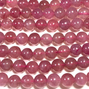 May include: Close-up of pink gemstone beads. The spherical beads vary in shade from pale to deep pink, with a translucent quality. They are strung together on a thin, pink thread, forming neat rows. The beads are likely for jewellery making.