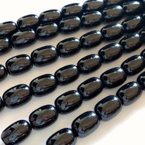 May include: Close-up of multiple strands of glossy, black, oval-shaped beads. The beads are uniform in size and shape, with a smooth, reflective surface. The beads are strung together, ready for jewelry making.