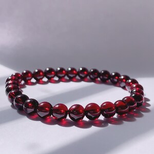 Puede incluir: Un brazalete de piedras preciosas rojas hecho de cuentas redondas. El brazalete est&aacute; sobre una superficie blanca.