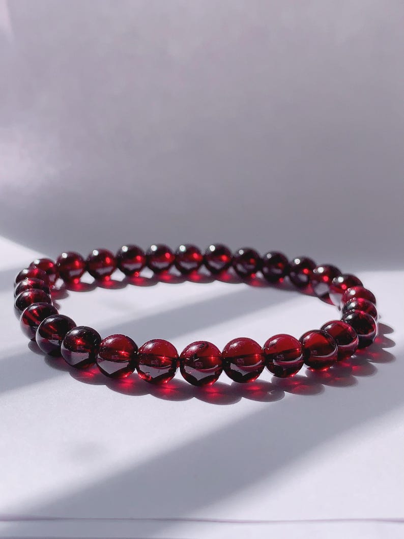 Puede incluir: Un brazalete de piedras preciosas rojas hecho de cuentas redondas. El brazalete est&aacute; sobre una superficie blanca.