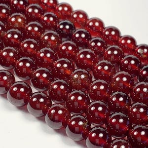 May include: Close-up of a strand of polished, round, dark red garnet beads. The beads have a glossy finish and reflect light, creating bright white spots. The beads are strung together, ready for jewellery making.