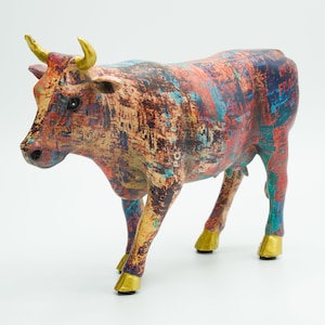 Peut inclure: Une figurine de vache en céramique colorée, avec un motif abstrait et des cornes et des sabots dorés. La vache est debout sur une surface blanche.