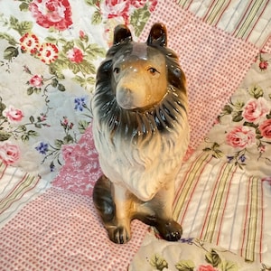Op de afbeelding: Een keramieken hondenbeeldje met een gedetailleerd, levensecht uiterlijk. De hond heeft een bruine en zwarte vacht met een witte borst en gezicht. Het beeldje staat op een quilt met bloemenpatroon.