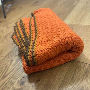 May include: Folded orange blanket with a textured surface. The blanket has a decorative border with brown, orange, and yellow stripes. The blanket is folded in half.