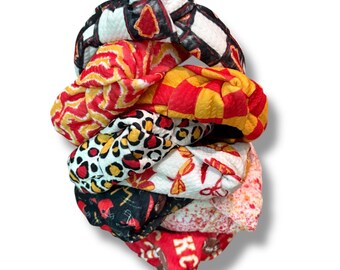 May include: A stack of knotted headbands in various patterns and colors. Designs include red and yellow geometric patterns, a black and white checkered pattern, and a leopard print. The headbands are made of fabric.