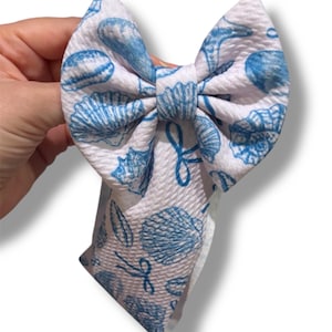 May include: A white fabric bow with a blue seashell pattern. The bow is held in a hand and has a textured surface. The bow is a hair accessory.